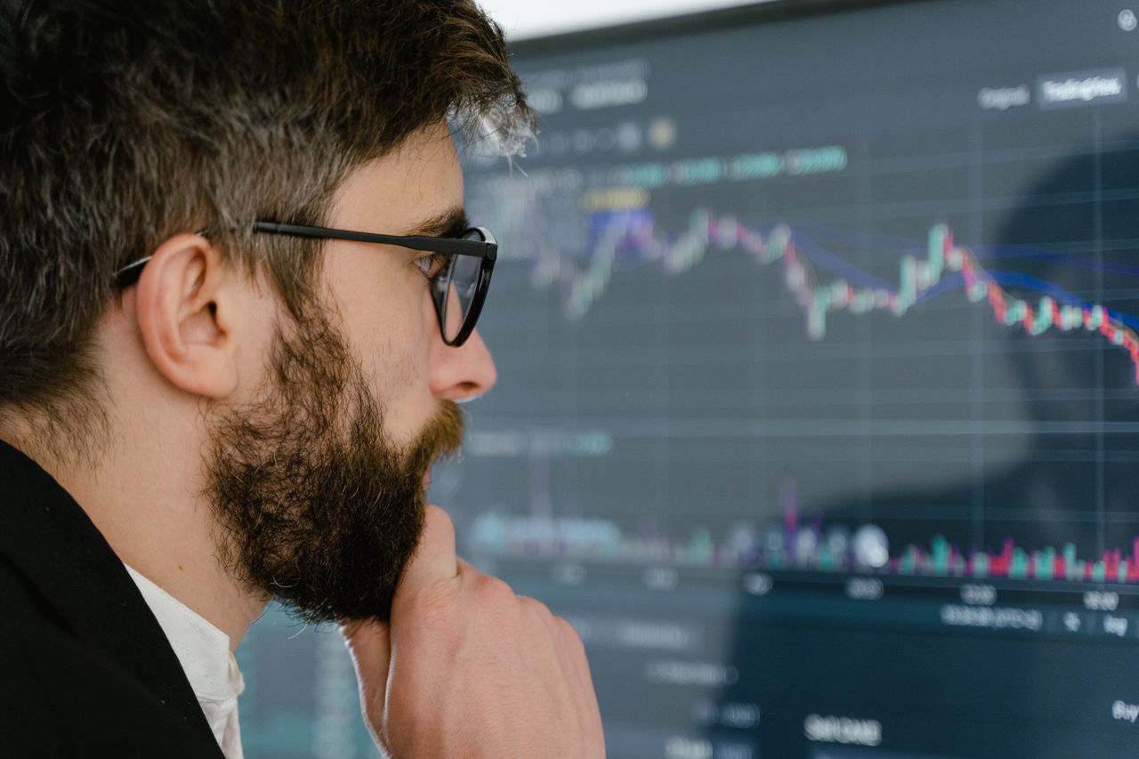 A focused man with glasses studies stock market graphs on a screen, pondering insights.